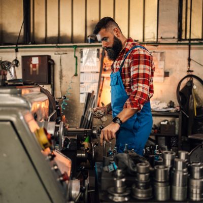 An image of a metal turner standing next to a lathe machine and a table with various metal pieces. He is operating the machine and holding a cloth. Small factory, workshop setting.Overalls and shirt.