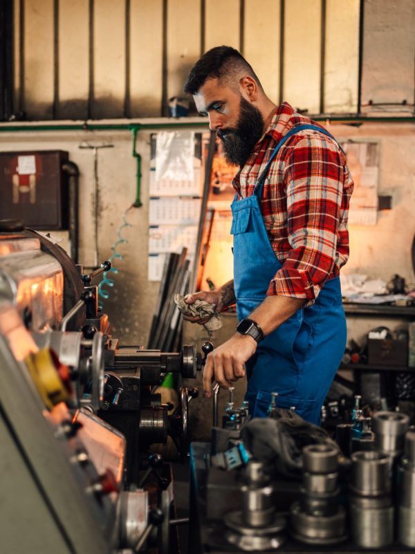 An image of a metal turner standing next to a lathe machine and a table with various metal pieces. He is operating the machine and holding a cloth. Small factory, workshop setting.Overalls and shirt.